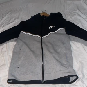 Grå och svart hoodie från Nike - Snygg och bekväm grå och svart hoodie från Nike med dragkedja och huva. Perfekt för en sportig look eller avslappnade dagar. Den har en vit Nike-logga på bröstet och kontrasterande detaljer.