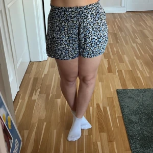Blommiga shorts från Divided H&M - Snygga blommiga shorts från Divided H&M i svart med blå och vita blommor. De har en elastisk midja för extra komfort och är perfekta för varma sommardagar. Materialet är mjukt och luftigt, vilket gör dem sköna att bära 🌟