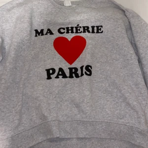 Grå sweatshirt med tryck - Säljer en grå sweatshirt med texten 'MA CHÉRIE PARIS' och ett rött hjärta på framsidan. Tröjan har en avslappnad passform och är perfekt för en casual look. Den är långärmad och gjord i mjukt material för extra komfort.