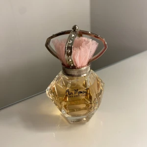 Our Moment parfym från One Direction - Elegant parfymflaska med en unik, facetterad design och en dekorativ rosa tyllprydd kork. Doften 'Our Moment' från One Direction är en fräsch och ungdomlig parfym, perfekt för fans av bandet eller de som söker en söt och blommig doft.