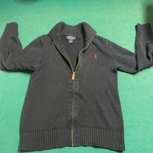 Svart kofta från Ralph Lauren - Säljer en stilren svart kofta från Ralph Lauren med dragkedja och ribbad krage. Den har det klassiska röda broderade logotypen på bröstet. Perfekt för en avslappnad men ändå elegant look.