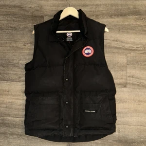 Svart dunväst från Canada Goose - Säljer en svart dunväst från Canada Goose med ett broderat märke på bröstet. Västen har knappar och praktiska fickor framtill. Perfekt för kyliga dagar.