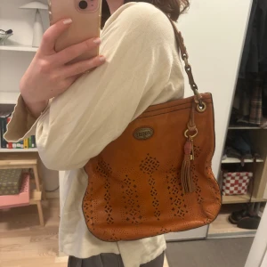 Brun axelväska från Fossil - Snygg brun axelväska från Fossil i skinn med dekorativa utskärningar och en tofsdetalj. Väskan har en axelrem och en dragkedja för säker förslutning. Perfekt för att ge din outfit en bohemisk touch.
