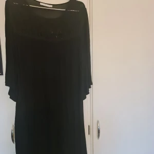 Svart klänning med spetsdetaljer - Elegant svart klänning med vackra spetsdetaljer vid halsringningen. Klänningen har en lös passform och är perfekt för en stilren look. Den är tillverkad i ett mjukt material som ger ett fint fall.