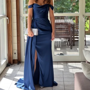 Marinblå långklänning från Dressroom Linné - Elegant marinblå långklänning från Dressroom Linné med offshoulder-design och en vacker slits framtill. Klänningen har en draperad detalj i midjan och ett släp som sitter fast vid sidan av midjan. Perfekt för speciella tillfällen. 
