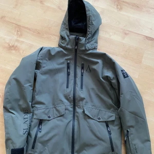 Grön jacka från Slade Couture - Snygg grön jacka från Slade Couture med huva och flera praktiska fickor med dragkedja. Perfekt för utomhusaktiviteter med sin funktionella design och stilrena look.