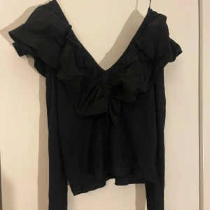 Svart blus med volang från Sybilla - Elegant svart blus från Sybilla med volangdetaljer och v-ringning. Blusen har långa ärmar och en stilren design som passar perfekt för olika tillfällen. Har aldrig kommit till användning men den passar också mig som brukar bära S bara att den kan va lite lösare eftersom den är i XL!
