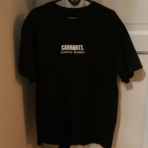 Svart t-shirt från Carhartt - Riktigt snygg och sällsynt svart t-shirt från Carhartt med tryck 'Synthetic Realities' på framsidan och ett coolt grafiskt motiv på baksidan. Perfekt för en avslappnad stil. Skriv gärna vid fler frågor!