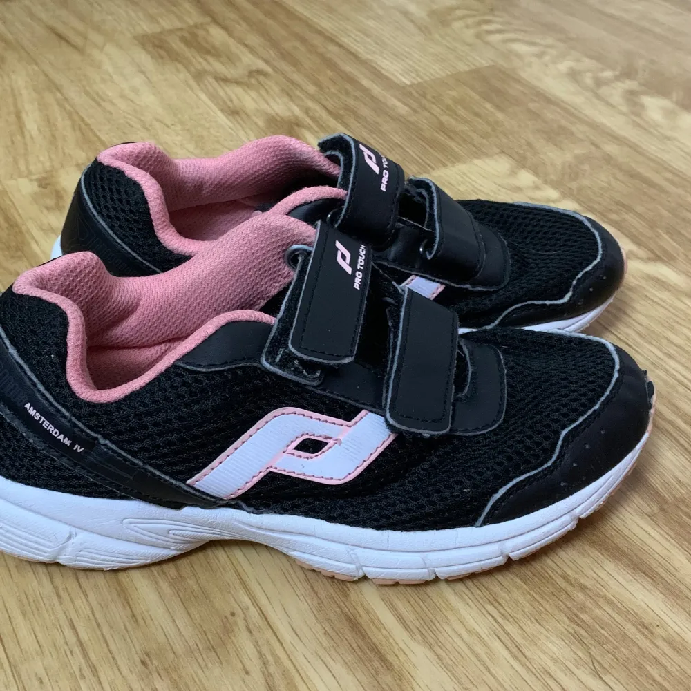 Säljer ett par snygga och bekväma sneakers från Pro Touch. Skorna är svarta med rosa detaljer och har en vit sula. De har två praktiska kardborreband för enkel på- och avtagning. Perfekta för både vardag och träning!. Skor.