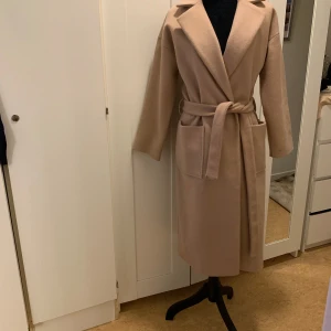 Beige kappa med bälte - Säljer en superfin beige kappa i nyskick. Den har en klassisk stil med ett bälte i midjan och långa ärmar. Perfekt för höst och vår när du vill se både stilren och mysig ut. Kappan är i en normal passform och passar till alla tillfällen.