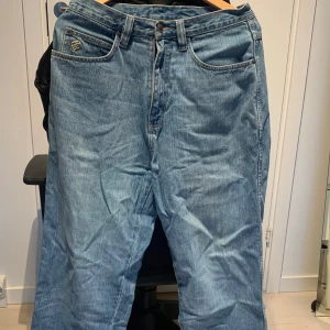 Blå jeans från Rocawear - Säljer ett par klassiska baggy blå jeans från Rocawear. De har en avslappnad passform med raka ben och är tillverkade i 100% bomull. Jeansen har en snygg broderad logga på bakfickan och en traditionell femficksdesign. Perfekta för en casual look!
