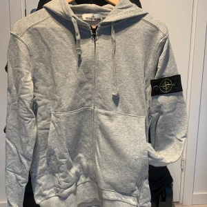 Grå hoodie från Stone Island - Säljer en snygg grå hoodie från Stone Island. Den har en dragkedja framtill och en klassisk huva med dragsko. På ärmen finns det ikoniska Stone Island-märket. Perfekt för en casual look eller när du vill hålla det stilrent och bekvämt.