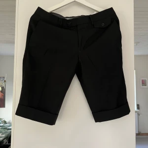 Svarta shorts från Neil Barrett - Säljer ett par stilrena svarta shorts från Neil Barrett, tillverkade i Italien. De är gjorda av 70% ull och 30% silke, vilket ger dem en lyxig känsla. Shortsen har en klassisk design med knappar och en snygg uppvikt kant. Perfekta för en elegant sommarlook!