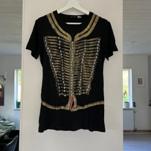 Svart topp med gulddetaljer från John Galliano - Säljer en snygg svart T-shirt från John Galliano, tillverkad i Italien. Den har korta ärmar och är dekorerad med guldfärgade detaljer och knappar framtill. Perfekt för en festlig kväll eller när du vill sticka ut lite extra!