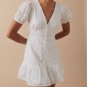 Vit klänning med spetsdetaljer - Säljer en vit klänning med spetsdetaljer och puffiga kortärmar. Den har en v-ringad design med knappar framtill och en volangkant längst ner. Storlek M från Ginatricot. Nyskick. Köpt för 499kr.