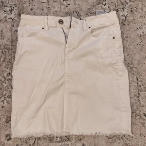 Beige jeanskjol med fransad kant - Säljer en superfin vit jeanskjol med fransad kant. Kjolen har en klassisk femficksdesign och stängs med knapp och dragkedja framtill. Perfekt för både vardag och festliga tillfällen. Passar bra till en avslappnad sommarlook!