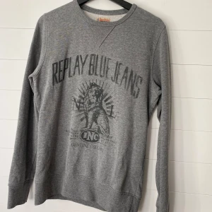 Grå tröja från Replay Blue Jeans - Säljer en snygg grå tröja från Replay Blue Jeans. Den har ett coolt tryck på framsidan och är perfekt för en avslappnad stil. Tröjan är långärmad och gjord i ett mjukt material som känns skönt mot huden. Perfekt för höst och vinter!