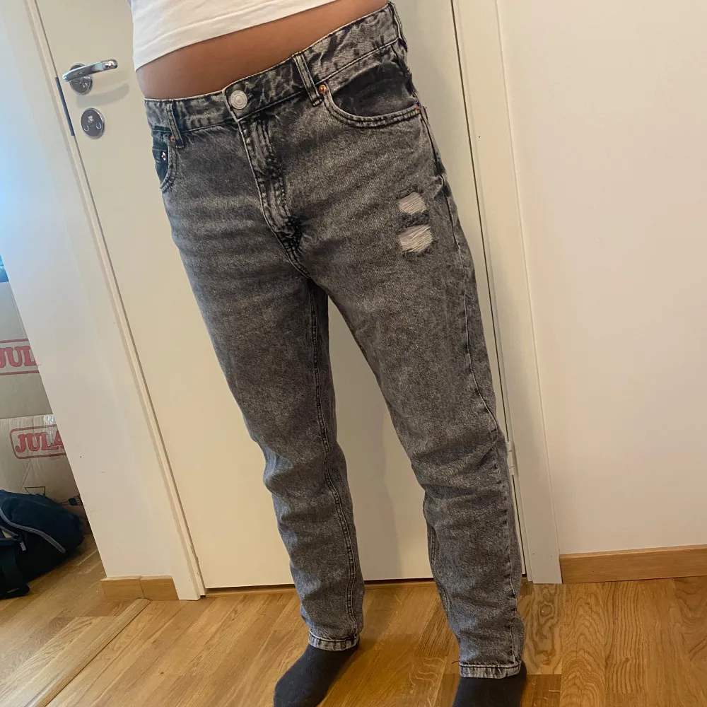 Säljer ett par grå jeans i nyskick med coola slitningar på framsidan. De har en klassisk femficksdesign och en knappgylf. Perfekta för en avslappnad stil och passar till det mesta. Storleken är normal och de är superbekväma att ha på sig.. Jeans & Byxor.