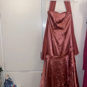 Rostfärgad axelbandslös klänning - Säljer en elegant rostfärgad axelbandslös klänning i satin. Den har en vacker volangdetalj och en smickrande passform som passar perfekt för festliga tillfällen. Klänningen är i nyskick och har en snörning  i ryggen
