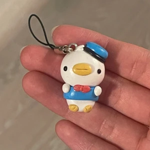 Söt anka nyckelring - En supergullig nyckelring med en liten anka klädd i blå sjömansdräkt och hatt. Ankan har en röd fluga och är perfekt som accessoar till väskan eller nycklarna. Den är liten och smidig, ungefär 4 cm lång, och har en svart rem för enkel fastsättning.
