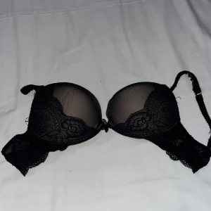 Svart spets-bh med rosett - Säljer en elegant svart bh med spetsdetaljer och en söt rosett framtill. Den har vadderade kupor och justerbara axelband för perfekt passform. Använd ett fåtal gånger, så den är i bra skick. 80D 
