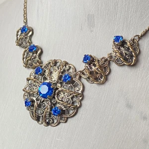 Vintage halsband med blå stenar - Säljer ett vackert vintage halsband med intrikata detaljer och gnistrande blå stenar. Halsbandet har en antik stil med ett stort, dekorativt hänge i mitten omgivet av mindre blå stenar. Perfekt för att ge en elegant touch till din outfit. Passar utmärkt till festliga tillfällen eller som en unik accessoar i vardagen.