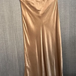 Champagnefärgad kjol - Elegant champagnefärgad kjol i ett glansigt material. Perfekt för festliga tillfällen med sin stilrena och sofistikerade look.och har en smickrande passform som framhäver figuren.