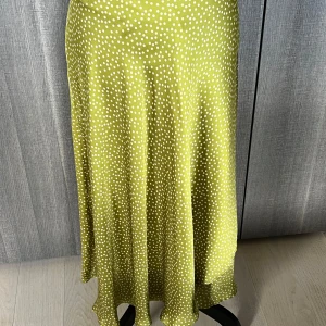 Grön prickig kjol - Säljer en superfin grön kjol med vita prickar. Kjolen är lång och har en härlig flowy känsla, perfekt för sommardagar eller en festlig kväll. Materialet känns som satin och ger en lyxig touch. Passar perfekt för dig som vill ha något unikt och stilrent i garderoben!