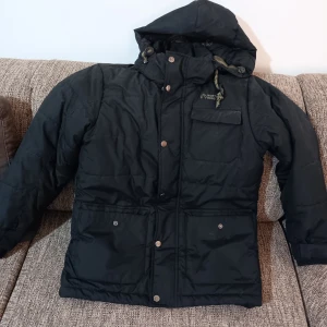 Svart vinterjacka från North Bend - Säljer en svart vinterjacka från North Bend i storlek M. Jackan har en avtagbar huva och stängs med både dragkedja och knappar. Den är perfekt för kalla vinterdagar och har flera praktiska fickor. Insidan är fodrad i en olivgrön nyans för extra värme. Perfekt för dig som vill hålla stilen i vinter! ❄️