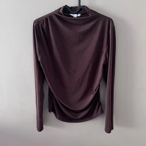 Brun långärmad topp - Säljer en elegant brun långärmad topp i mjukt material. Toppen har en draperad design som ger en snygg och stilren look. Perfekt för höst och vinter, både till vardags och fest. Passar bra med jeans eller kjol för en chic outfit.