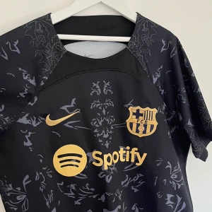 Svart FC Barcelona fotbollströja - Säljer en svart FC Barcelona fotbollströja från Nike med guldiga detaljer och mönster. Tröjan har korta ärmar och är i nyskick. Perfekt för alla Barca-fans som vill ha en stilren look. Passar både till match och vardag!
