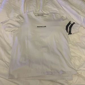 Vit t-shirt från Moncler - Säljer en snygg vit t-shirt från Moncler i storlek M. Den har en klassisk design med Moncler-loggan tryckt på bröstet och en cool detalj på ärmen. Perfekt för en stilren och avslappnad look. Passar både till vardags och fest!