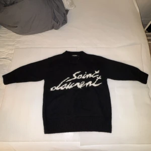 Svart stickad tröja från Saint Laurent - Snygg svart stickad tröja från Saint Laurent med deras ikoniska logga i vitt på framsidan. Tröjan har långa ärmar och en ribbad kant vid halsen, ärmsluten och nederkanten. Perfekt för en stilren look.