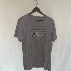Grå t-shirt från Ralph Lauren - Snygg grå t-shirt från Ralph Lauren med tryck 'Ralph 67' på framsidan. Perfekt för en avslappnad stil. T-shirten har en klassisk passform och är kortärmad.L passar mer M 