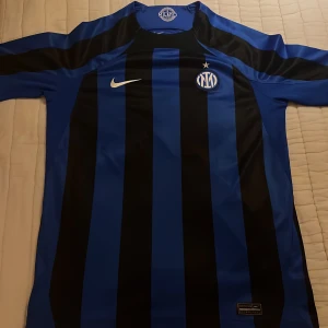 Blå och svart randig fotbollströja från Nike - Säljer en blå och svart randig fotbollströja från Nike med Inter-logga på bröstet och Lenovo-tryck på ryggen. Tröjan är kortärmad och tillverkad i Dri-FIT-material för optimal komfort. Perfekt för fotbollsfans som vill visa sitt stöd för laget.