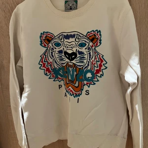 Vit sweatshirt från Kenzo med tigerbroderi - Säljer en vit sweatshirt från Kenzo med ett stort, färgglatt tigerbroderi på framsidan. Tröjan har rund hals och långa ärmar, perfekt för en stilren look med en touch av färg. Broderiet har detaljer i blått, rött och orange.