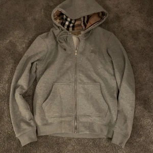 Grå hoodie från Burberry - Snygg grå hoodie från Burberry med dragkedja och klassiskt rutigt foder i huvan. Perfekt för en stilren och bekväm look. Den har långa ärmar och praktiska fickor framtill.