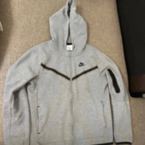 Grå nike tech - Säljer en grå hoodie från Nike med dragkedja och huva. Den har en svart randdetalj över bröstet och en ficka på ärmen. Perfekt för en sportig look! Bra skick storlek 147-158