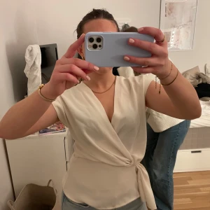 Vit omlottopp från Zara - Säljer en stilren vit omlottopp från Zara. Toppen har en knytning i midjan och korta ärmar, vilket ger en elegant look. Perfekt för både vardag och festliga tillfällen.