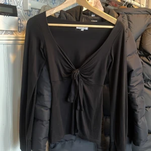 Svart långärmad topp från Design by Si - Säljer en stilren svart långärmad topp från Design Si. Toppen har en knytning framtill som ger en fin detalj och en v-ringad design. Perfekt för att skapa en elegant look.
