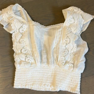 Vit blus från Pimkie - Charmig vit blus från Pimkie med spetsdetaljer och smockad nederkant. Blusen har korta ärmar och är perfekt för en somrig look!