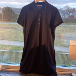 Mörkblå klänning från Lacoste - Säljer en mörkblå klänning från Lacoste i slim fit. Klänningen har korta ärmar och knappar framtill. Perfekt för en avslappnad men ändå elegant look. 🐊