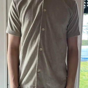 Beige kortärmad skjorta - Säljer en stilren beige kortärmad skjorta med knappar. Perfekt för en avslappnad look. Skjortan har en normal passform och är tillverkad i ett Frotté material. Väldigt luftig och går att ha till nästan allting. Vid några frågor är det bara höra av sig!💪