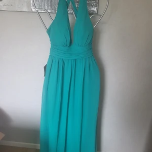 Turkos långklänning - Snygg turkos långklänning med halterneck och korsade band i ryggen. Perfekt för sommarens fester med sin eleganta och luftiga design. Klänningen har en vacker drapering vid midjan som ger en smickrande passform.