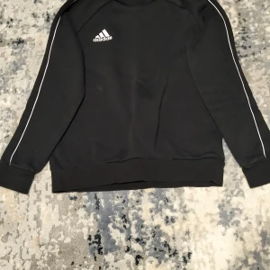 Svart tröja från Adidas - Säljer en svart tröja från Adidas med vita ränder längs ärmarna och logotyp på bröstet. Tröjan har långa ärmar och en klassisk passform. Perfekt för en sportig look eller avslappnad stil.