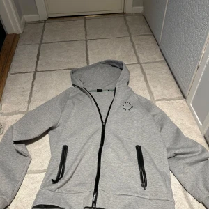 Boss hoodie i grå färg - Säljer en stilren grå hoodie från Boss med dragkedja och svarta detaljer. Köpte den på johnells förra sommaren och bara använd ca 3-4 gånger sedan dess. 
