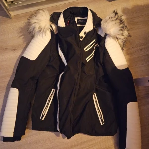 Svart och vit jacka - Snygg svart och vit jacka från John med pälsdetaljer på huvan. Jackan har flera dragkedjor och quiltade partier på ärmarna. Perfekt för kyligare dagar.