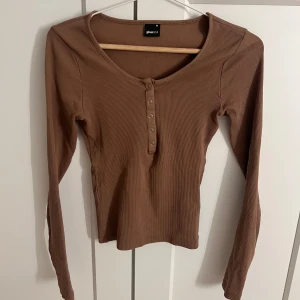 Brun ribbad långärmad topp från Gina - Säljer en snygg brun ribbad topp från Gina med långa ärmar och knappar framtill. Perfekt för en stilren look. Passar bra till både jeans och kjol. Hör av dig vid intresse!