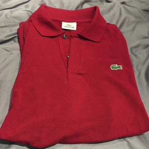 Röd pikétröja från Lacoste - Snygg röd pikétröja från Lacoste med klassisk krage och knappar framtill. Tröjan har det ikoniska gröna krokodilemblem på bröstet. Perfekt för en stilren och avslappnad look.