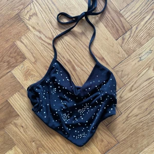 Svart halterneck topp med pärlor - Snygg svart halterneck topp med pärldetaljer. Perfekt för fest eller varmare väder. Toppen har en v-ringad design och knytband runt halsen.